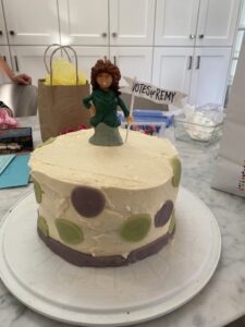 Round cake with white frosting and purple and green polka dots. A marzapan suffragette figurine is on top with a flag that reads Votes for Remy