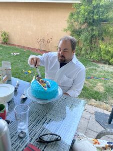 Mark sitting in a white shirt in front of the blue cake getting ready to cut it